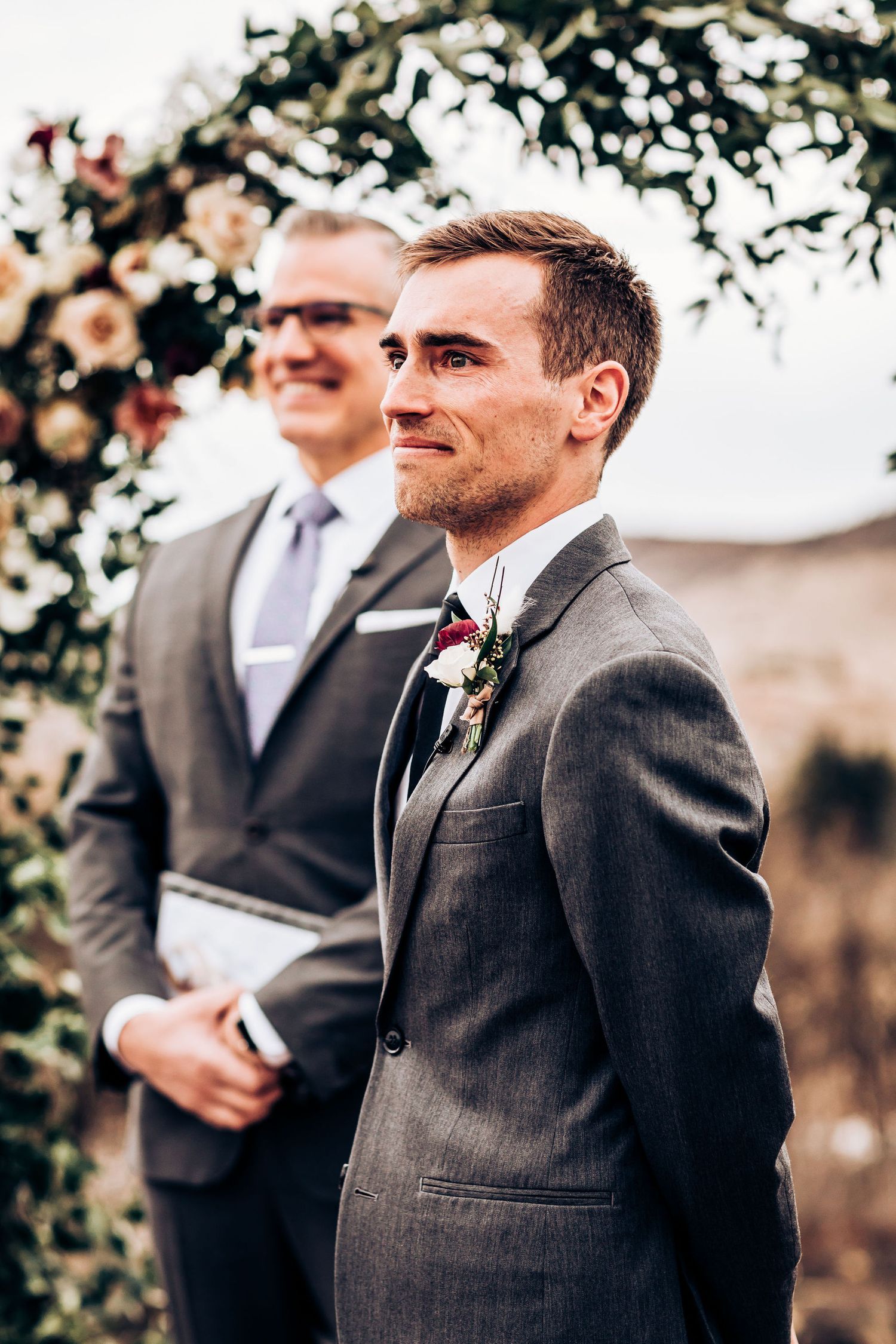 Men in formal grey suits stand beneath floral wedding arch.