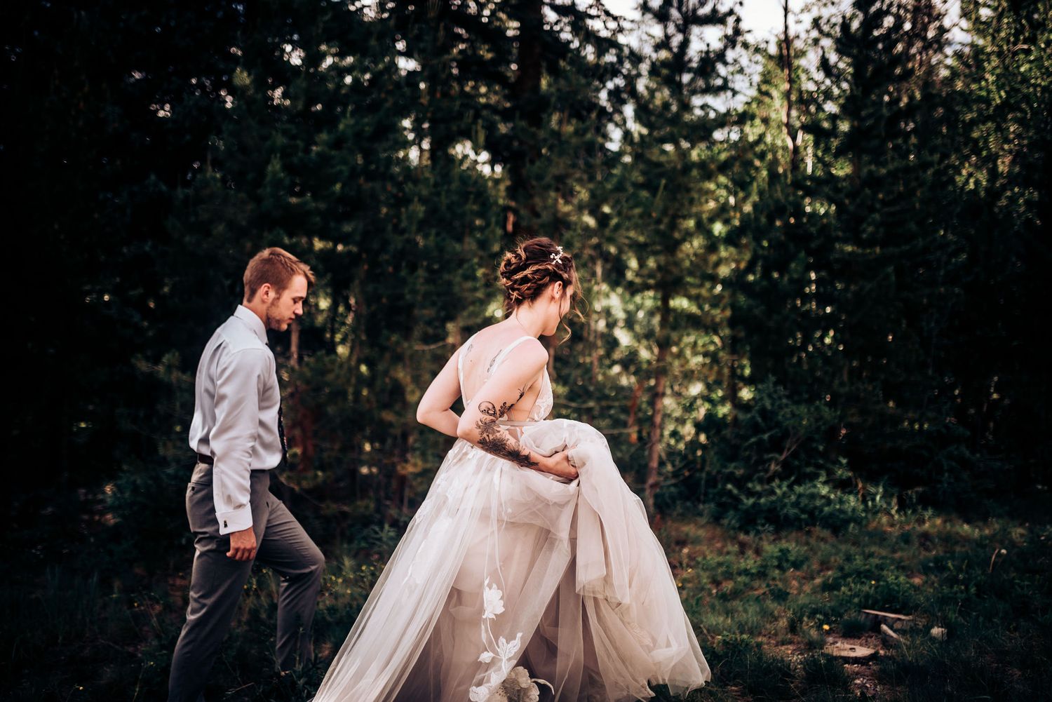 A couple walks through a wooded area, with the bride lifting her flowing wedding dress.