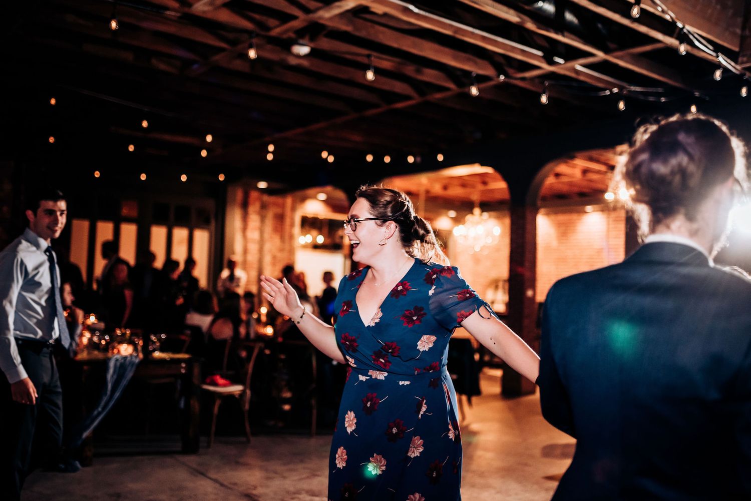 Wedding guests dance and celebrate at rustic venue with exposed brick and string lights.