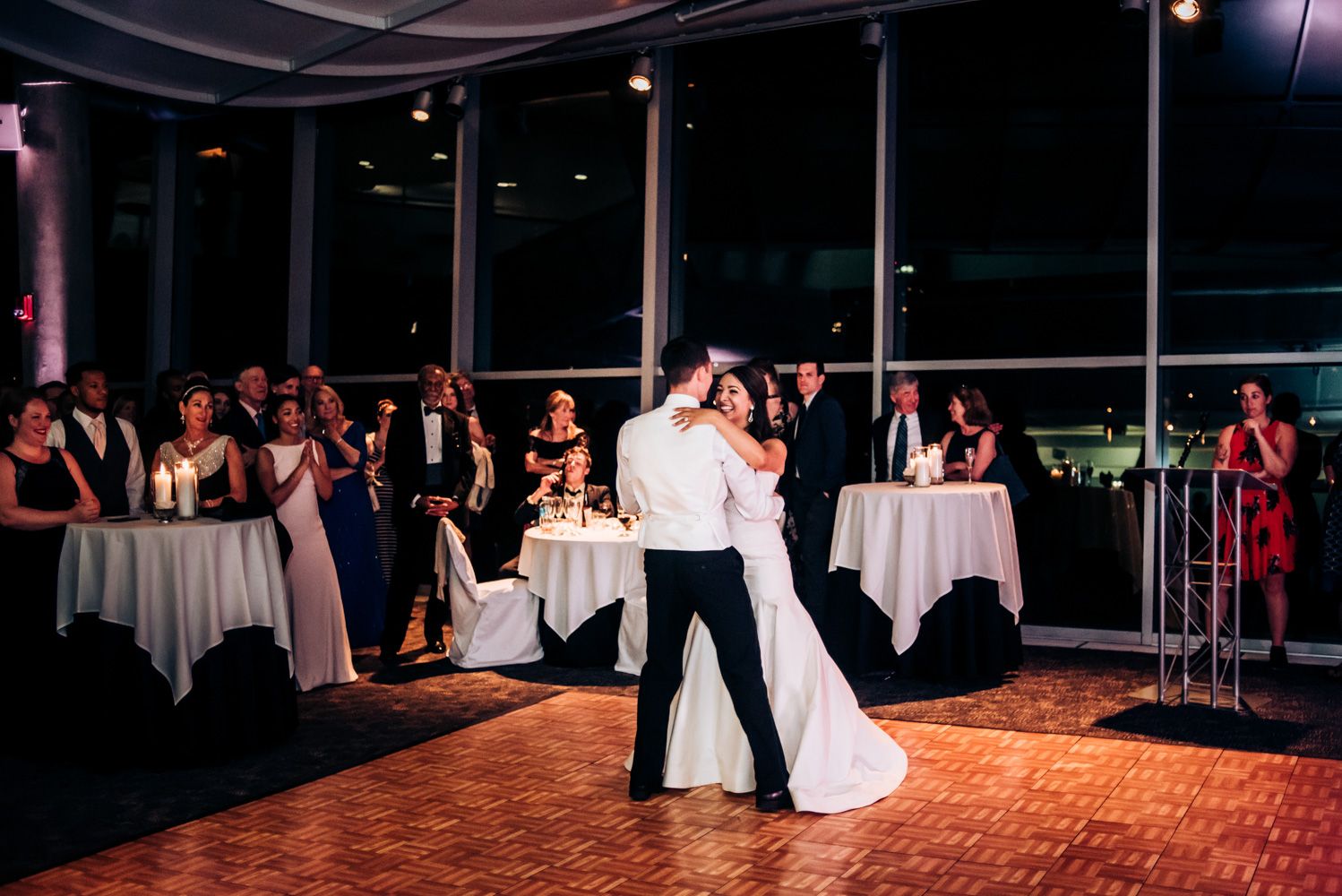 Newlyweds dance together on a wooden floor while wedding guests watch from surrounding tables.