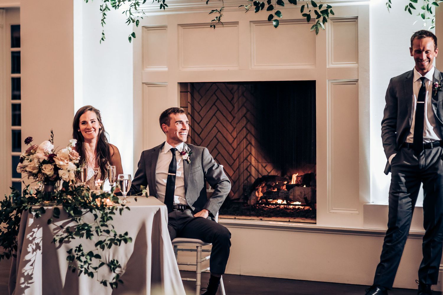 Wedding party members seated at decorated head table near elegant fireplace with floral arrangements.