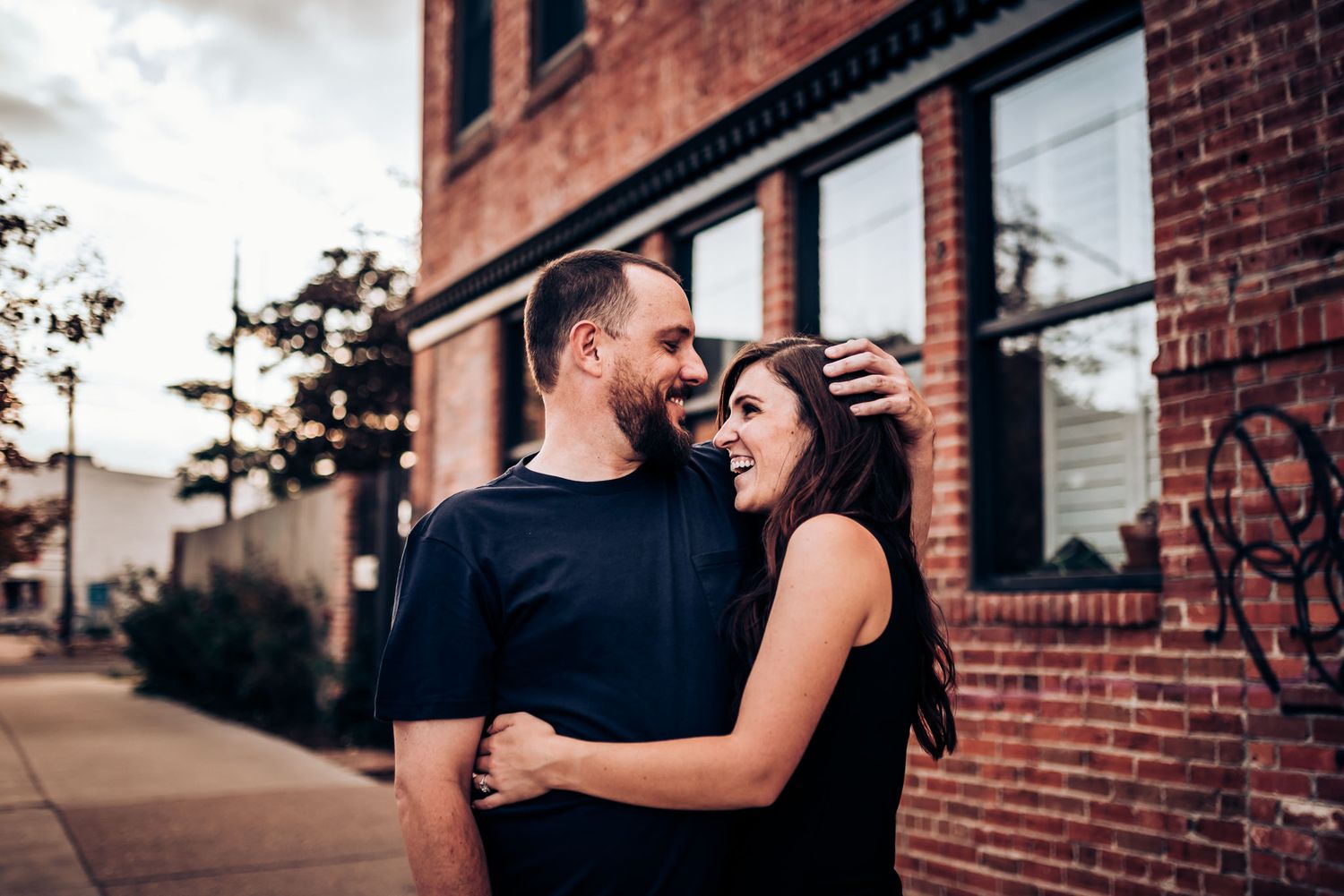 A couple shares an intimate moment outside a brick building with large windows.