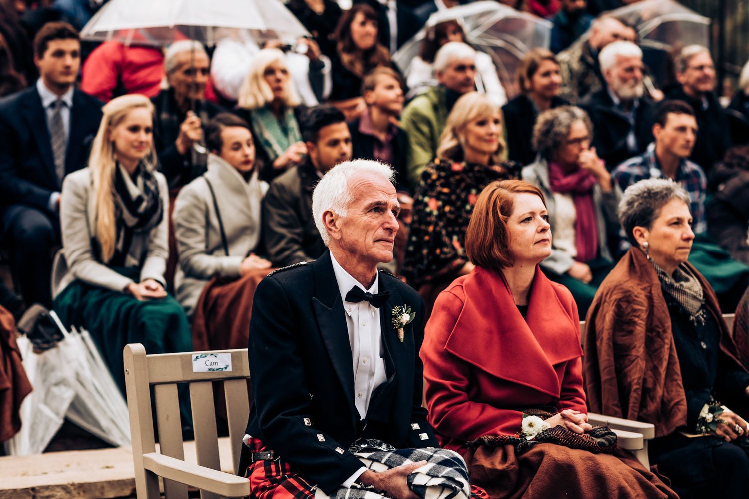 Wedding guests seated outdoors in formal attire on wooden chairs during an autumn ceremony.