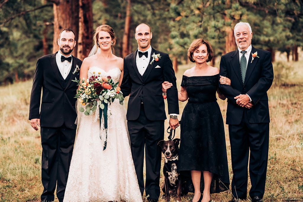 Wedding party in formal attire poses with black and white dog in outdoor setting.