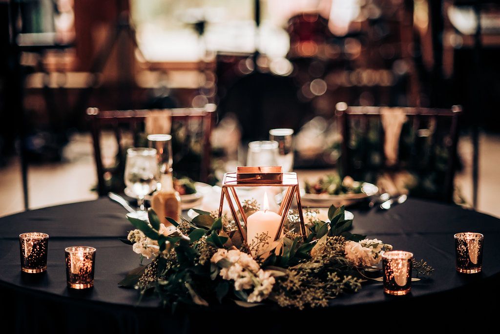 Romantic wedding centerpiece with glowing candles and greenery on dark tablecloth.
