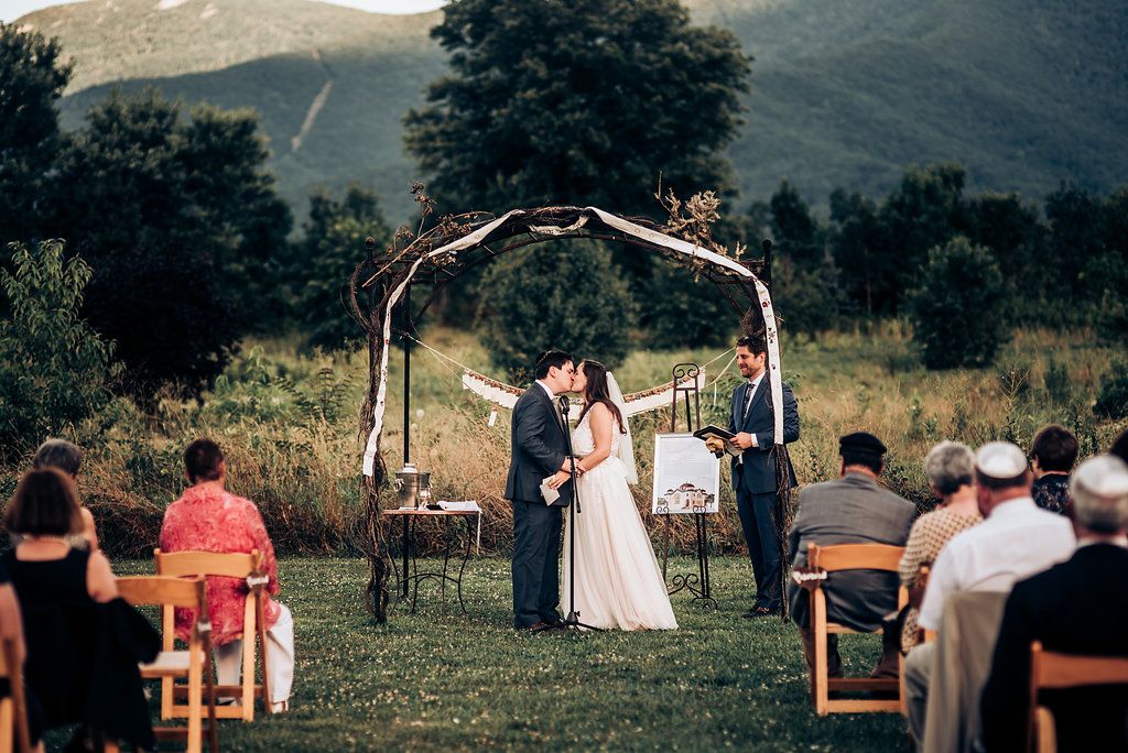 Outdoor wedding ceremony beneath rustic arch against mountain backdrop with seated guests.