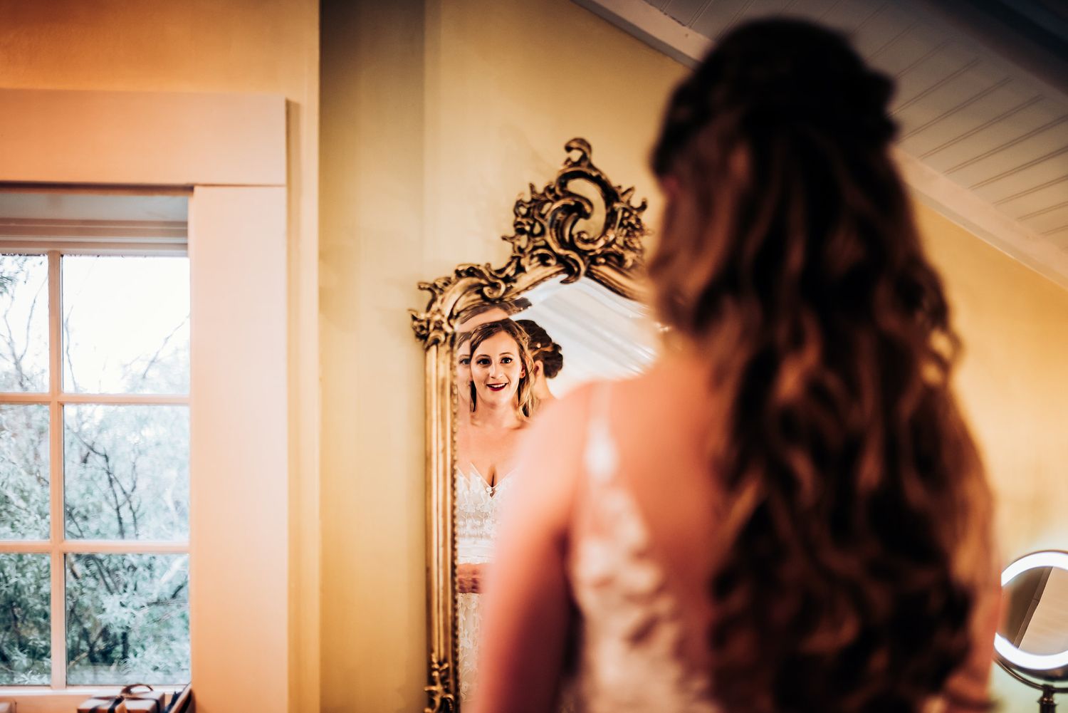 Reflection in ornate gold mirror during wedding preparations in sunlit room.