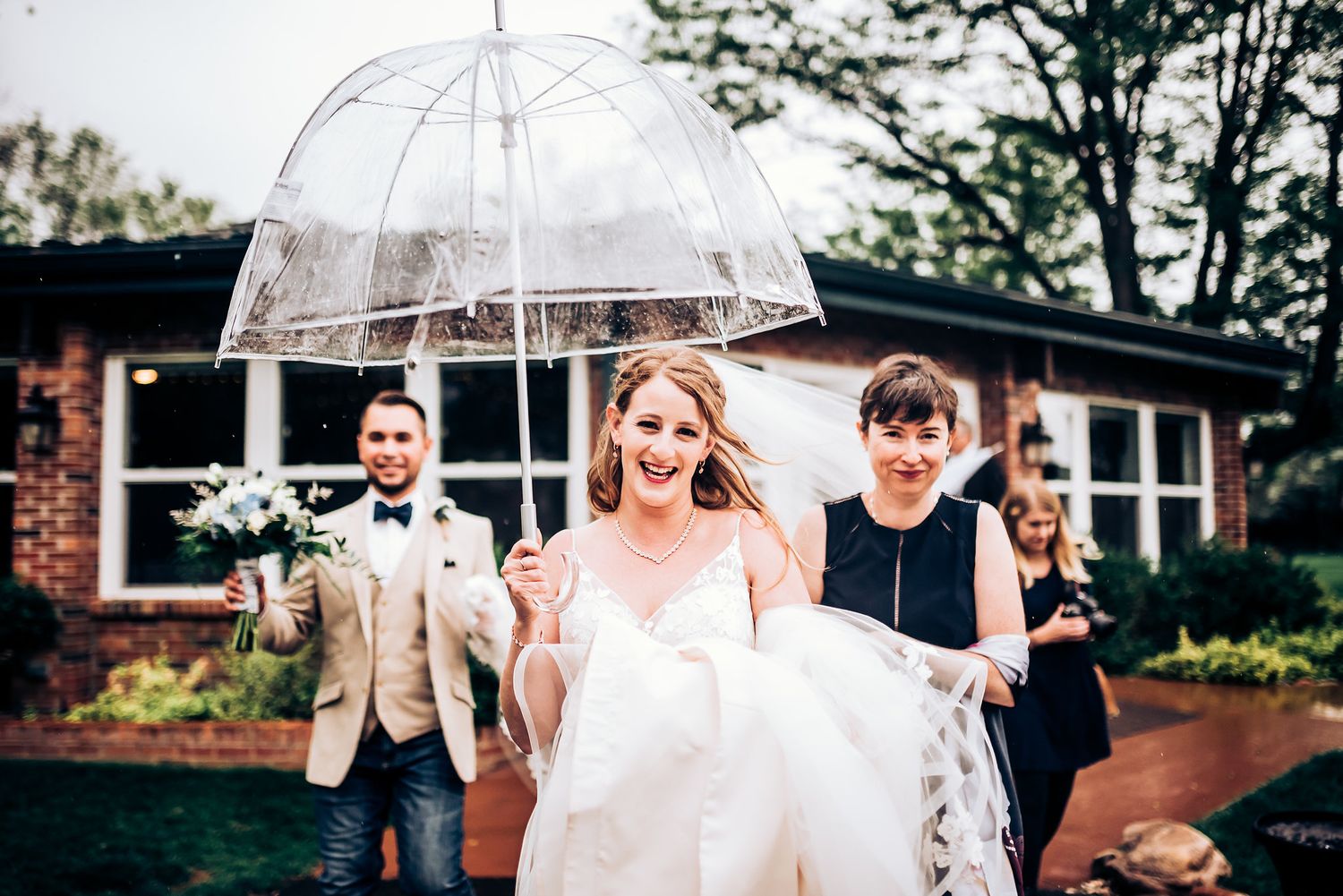 Joyful wedding scene with clear umbrella and brick building backdrop.
