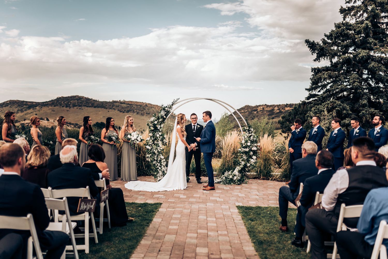 Panoramic view of outdoor wedding ceremony with guests seated on white chairs facing decorated arch.