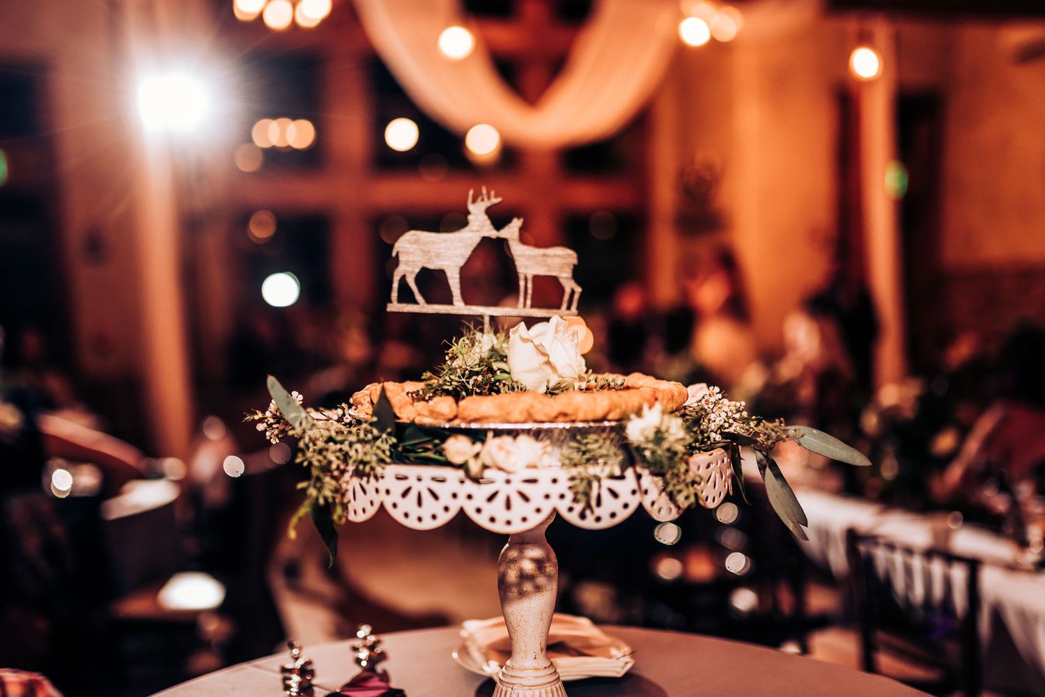 Elegant wedding cake topped with deer figurines displayed on ornate lace stand at reception.