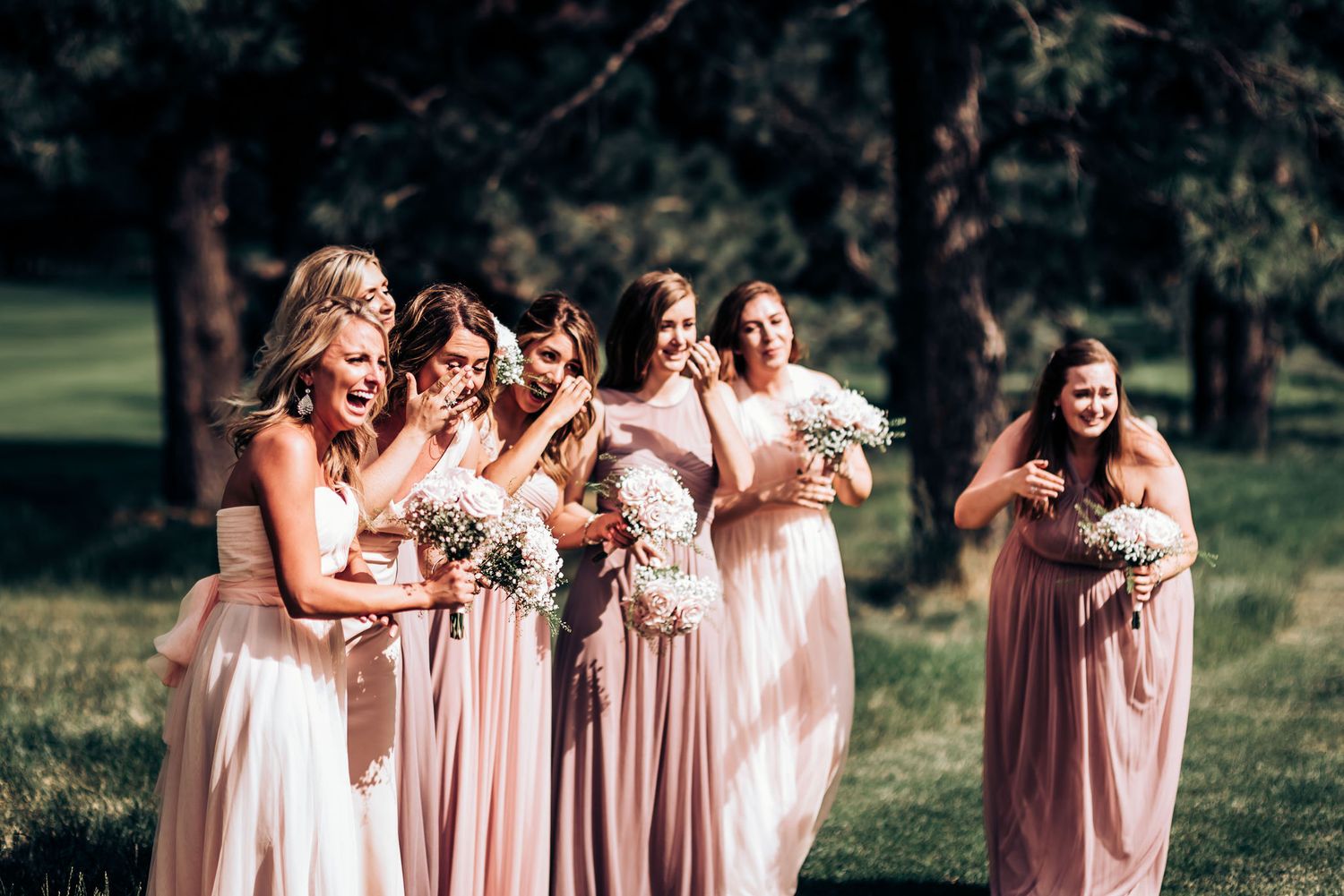 Wedding party in flowing pink dresses share candid laughing moment on lawn.