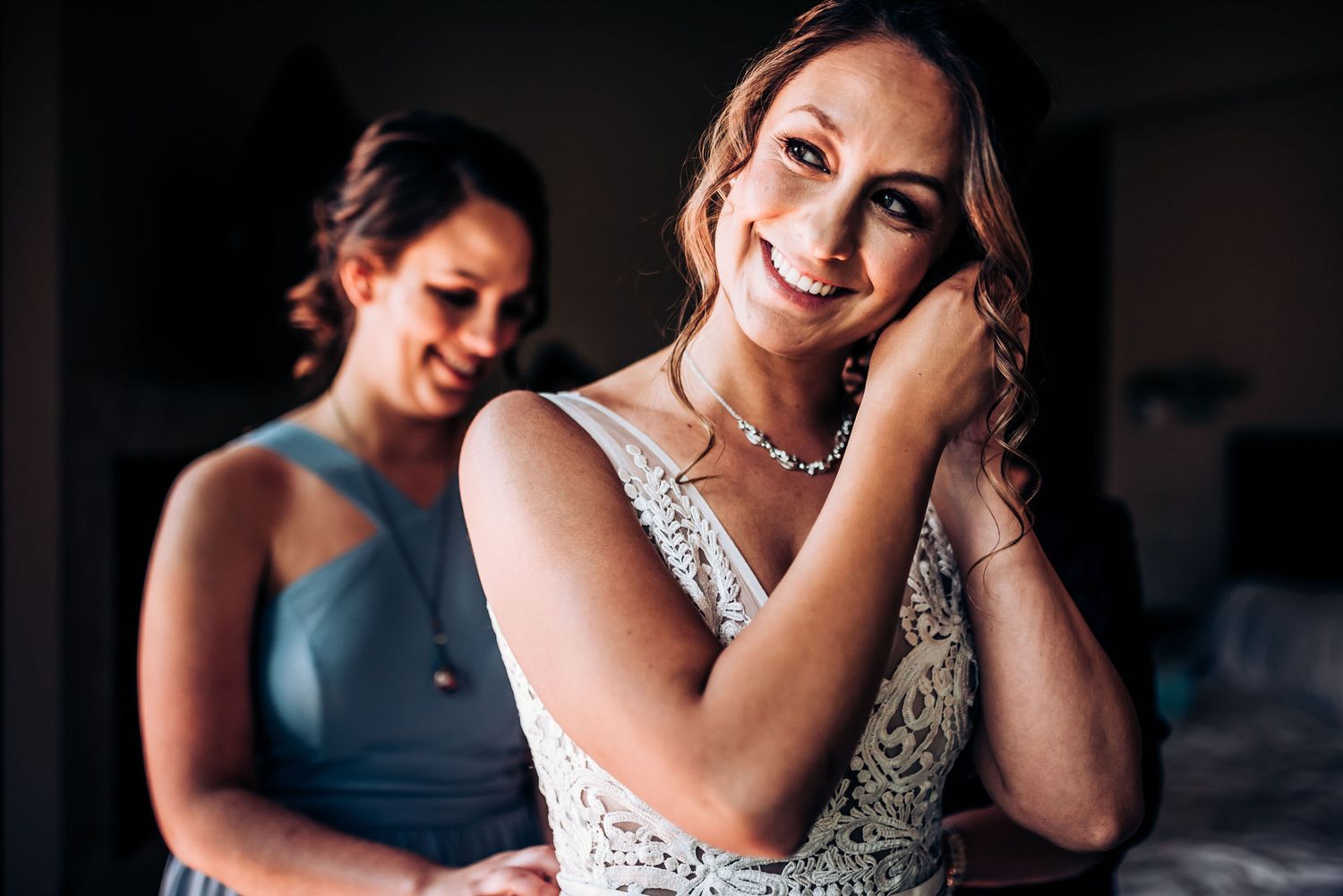 Bride in lace wedding gown touches necklace while smiling radiantly.