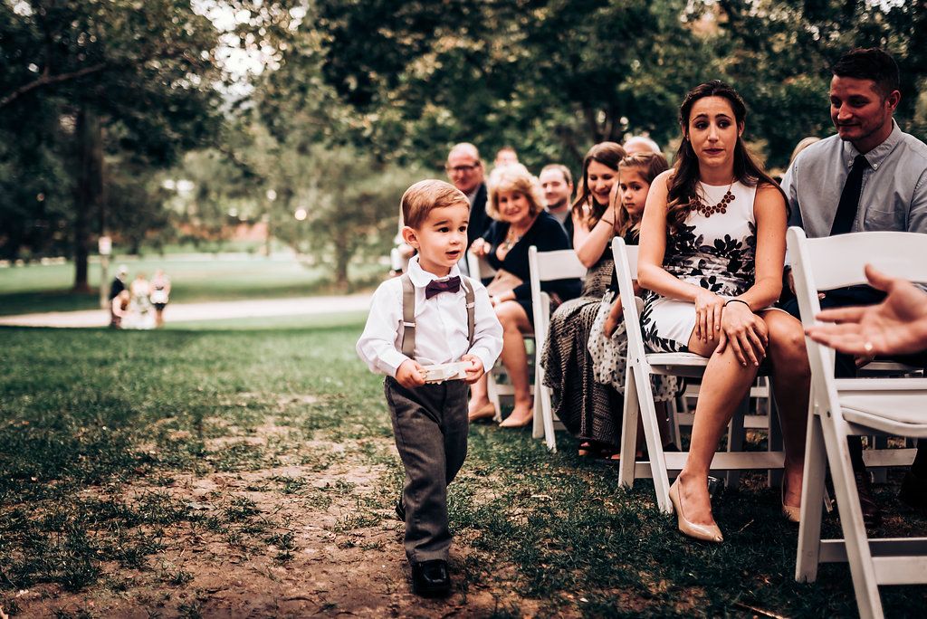 Young attendant in suspenders walks past seated wedding guests on outdoor grass aisle.