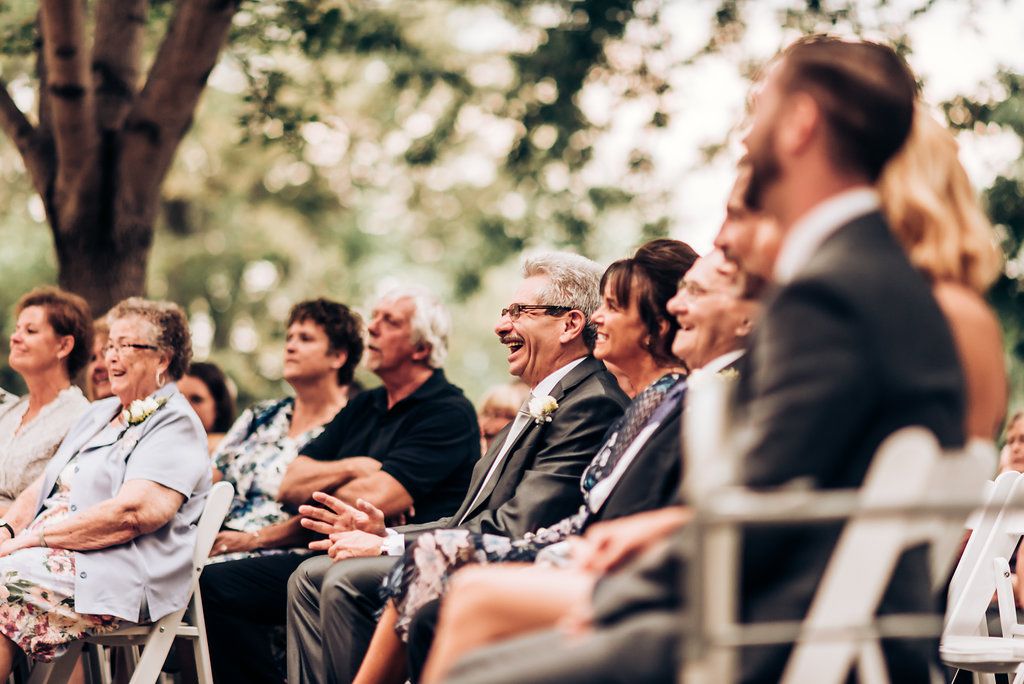 Wedding guests seated in white chairs watch the ceremony in a garden setting.