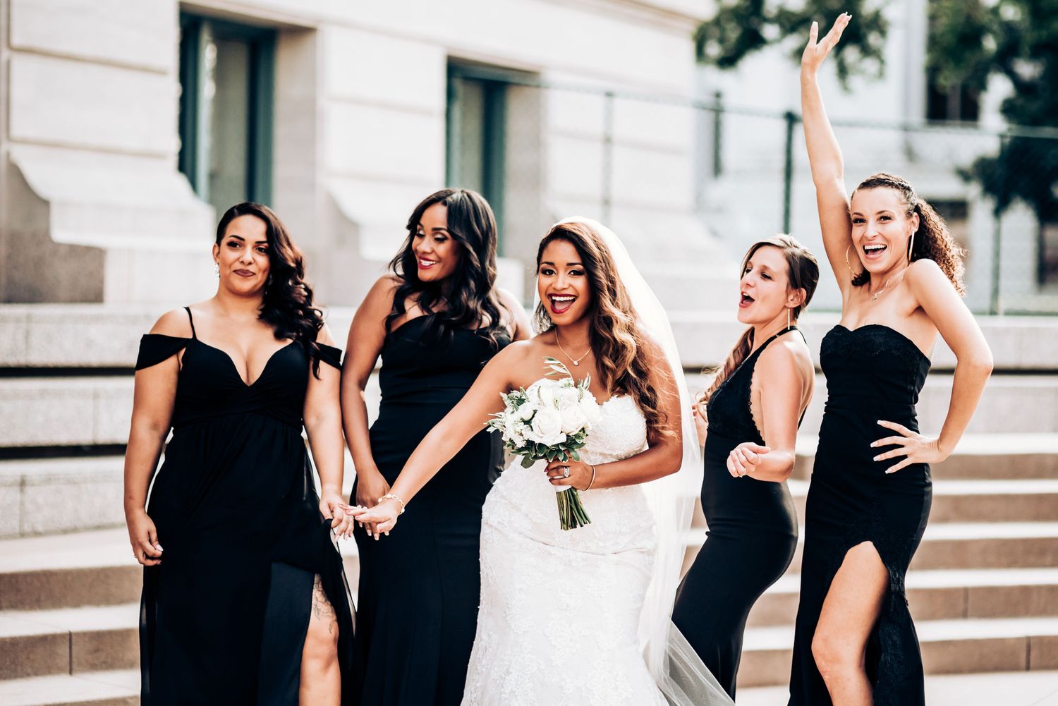 Wedding party wearing elegant black dresses poses with bride in white gown holding bouquet on stone steps.
