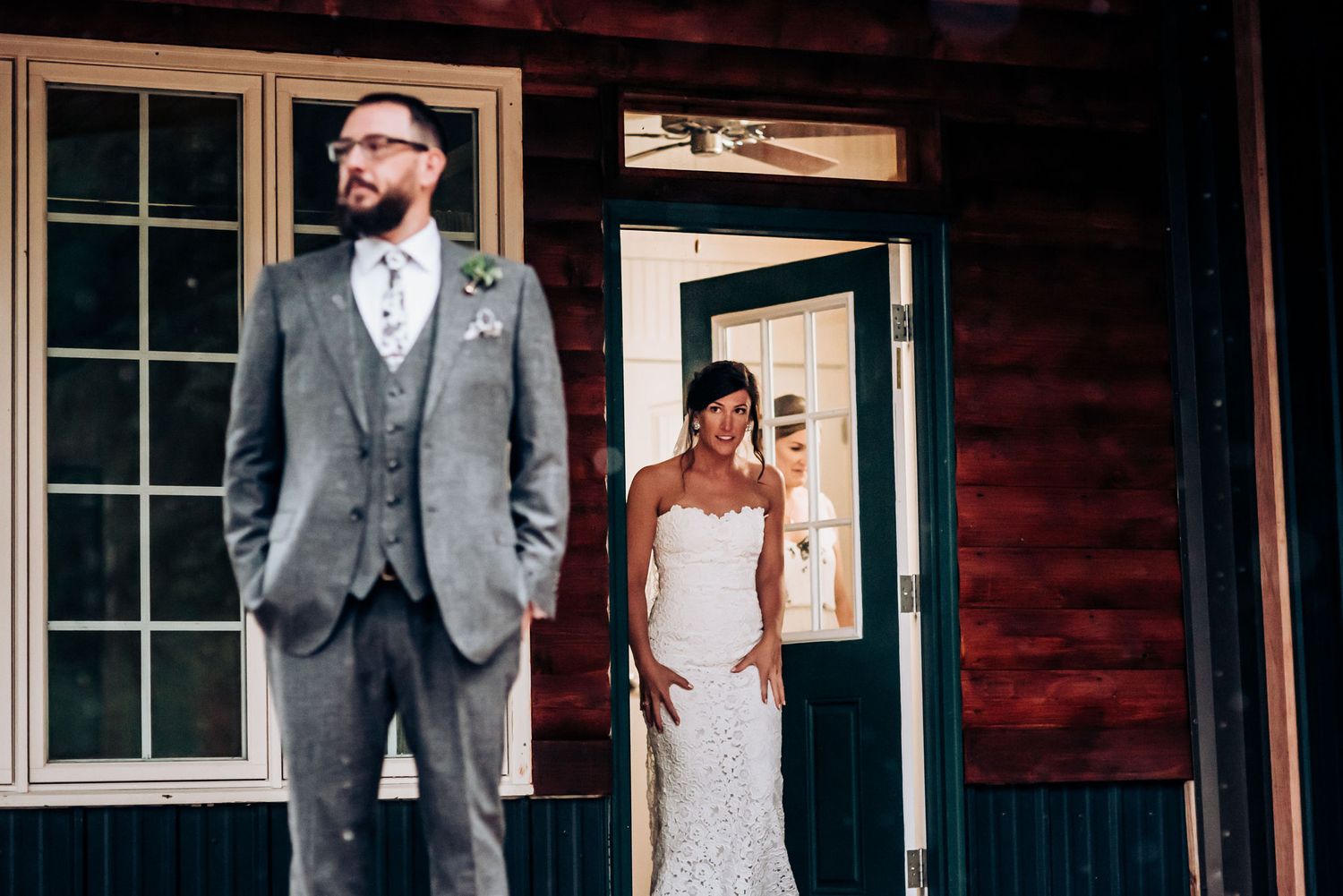 Wedding couple poses separately near rustic wooden cabin with large windows.