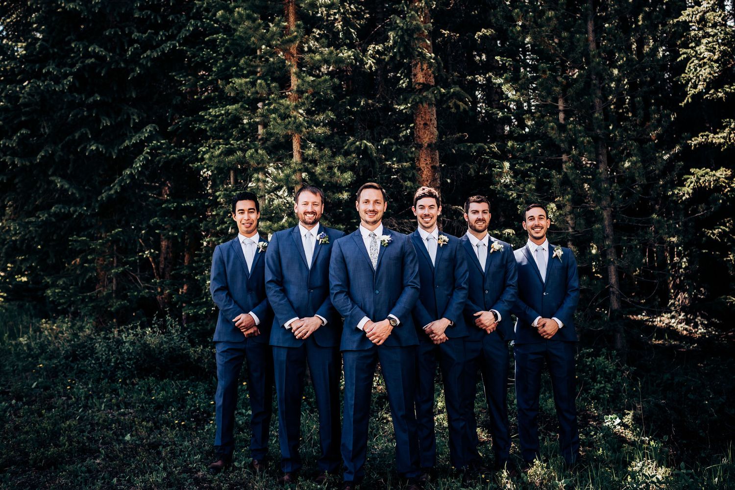 Groomsmen in matching navy blue suits pose together in a forest setting.