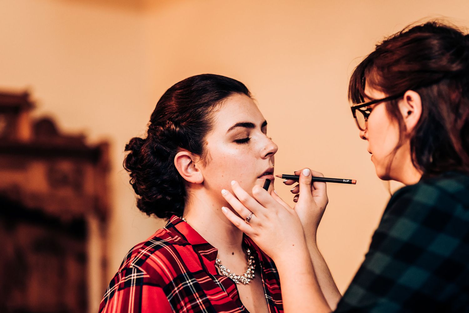 Makeup artist applying cosmetics in a warm-toned setting with red plaid accents.