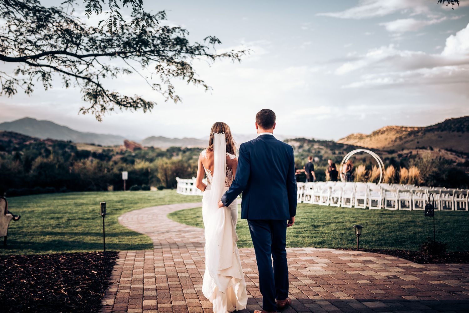 Newlyweds walk along a brick path overlooking a mountain landscape with white chairs set up for ceremony.