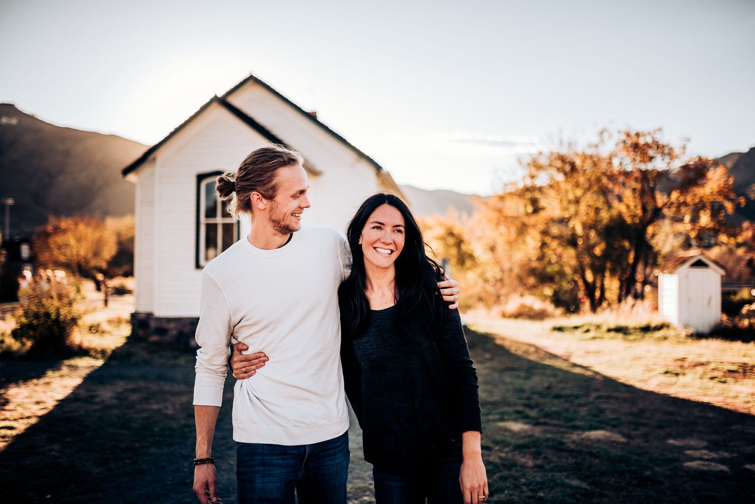 A couple stands together in front of a white house during golden hour in a rustic mountain setting.