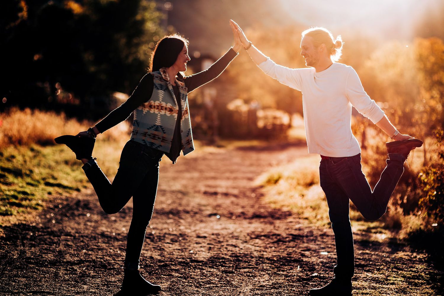 Playful couple high-fiving while balancing on one leg at sunset.