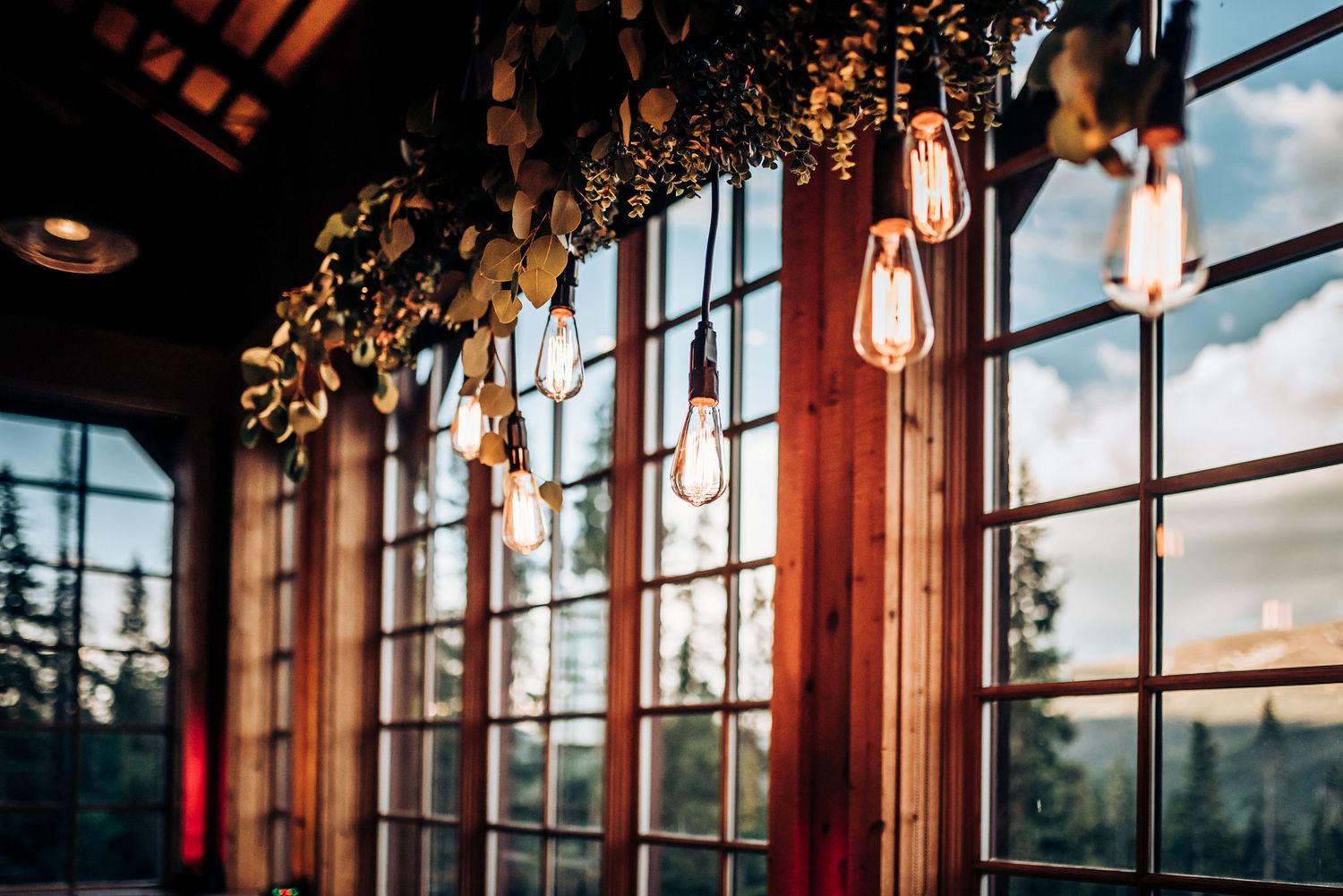 Large windows frame mountain sunset view with hanging Edison bulbs and greenery.