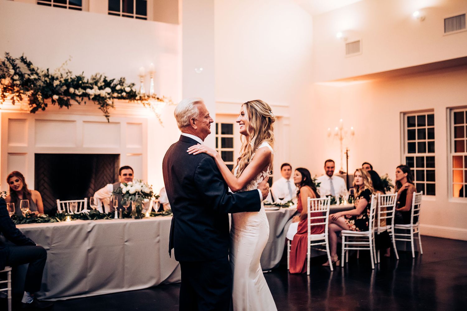 A couple shares their first dance in an elegant reception hall with white decor and flowers.
