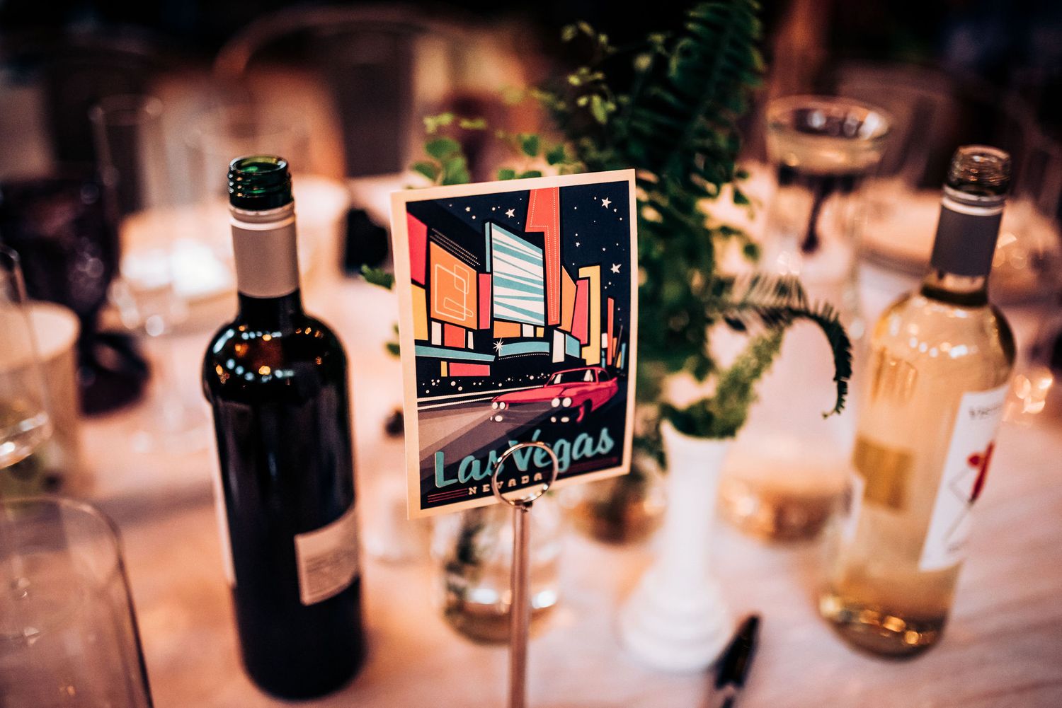 Vintage travel poster table markers with wine bottles and greenery at wedding reception.