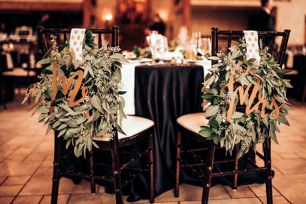 Wedding reception chairs decorated with cascading greenery garlands and white ribbons around black tablecloth.