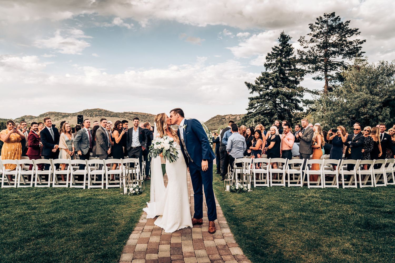Wedding ceremony on stone path with evergreen trees and rolling hills in background.