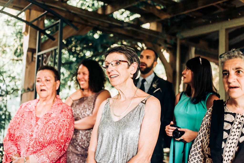 Wedding guests in colorful attire gather under a rustic wooden pergola during the ceremony.