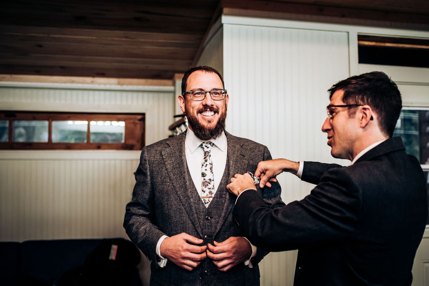 Two men in formal attire help adjust a tie and share a laugh during wedding preparations.