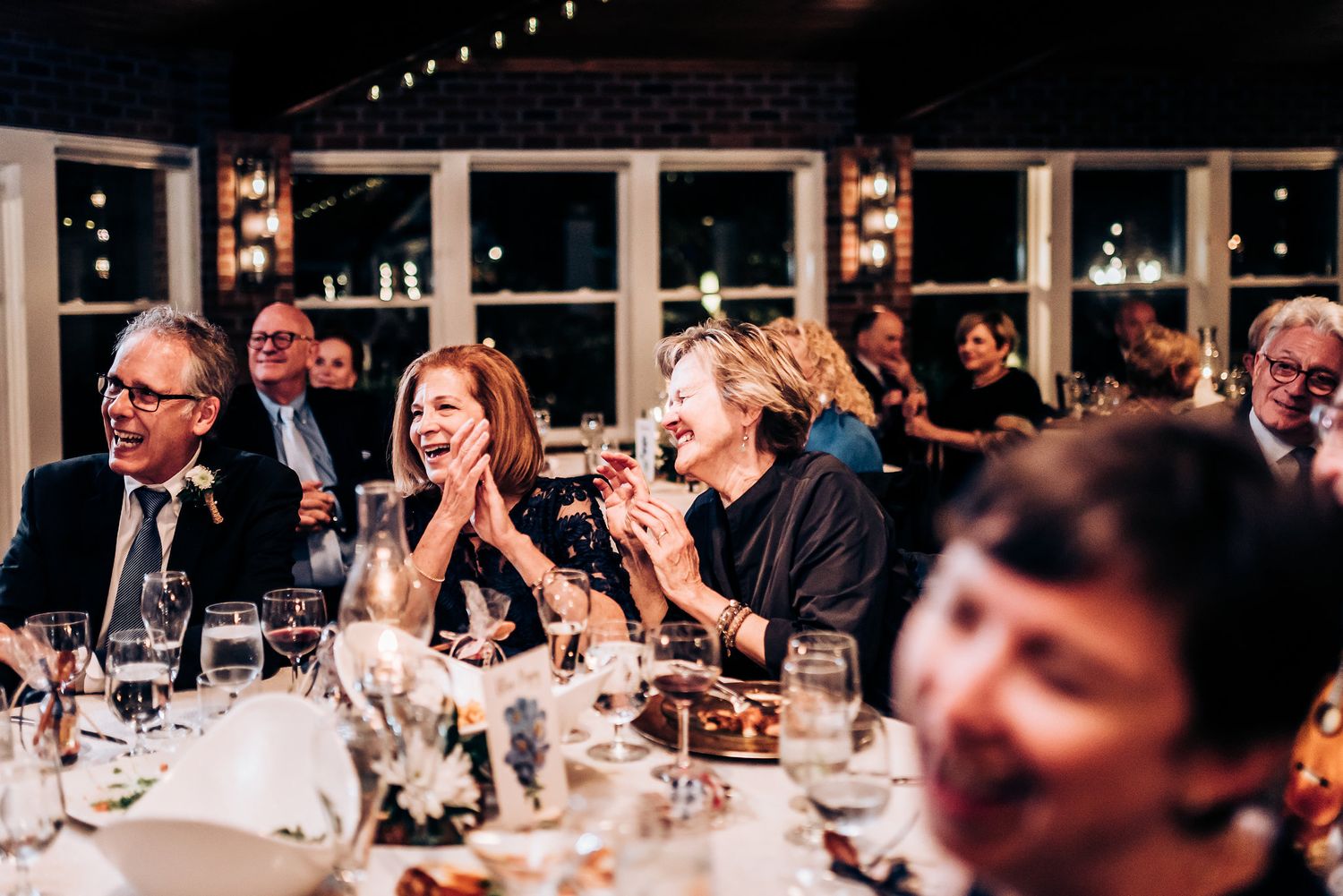 Wedding reception guests laugh and celebrate during dinner at tables with string lights overhead.