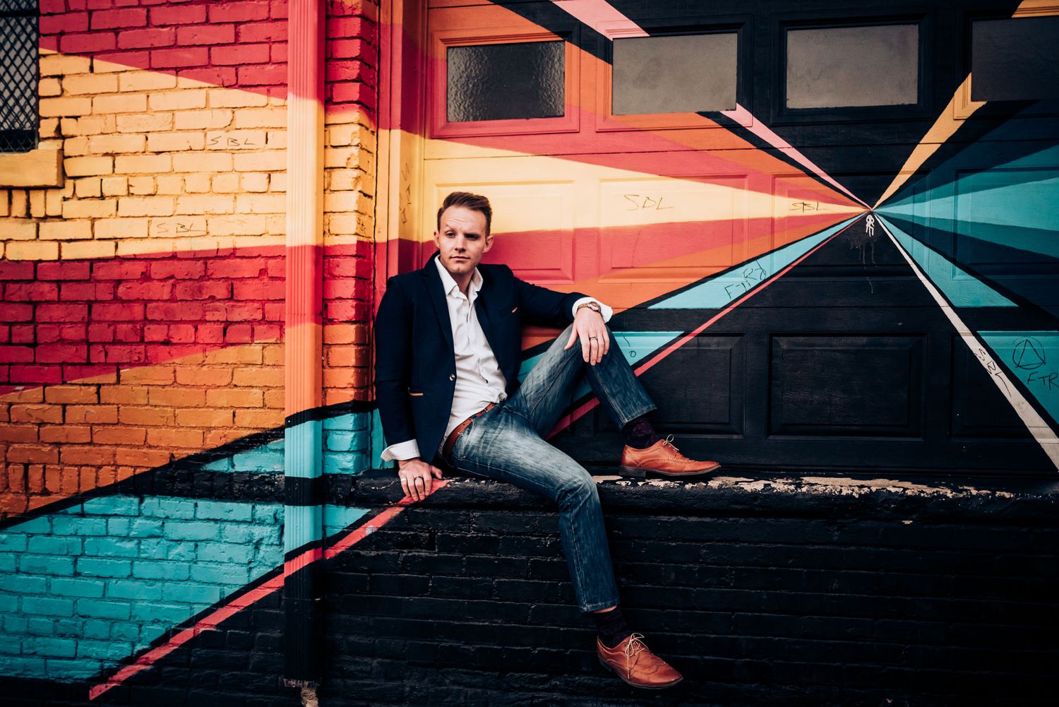 Person in dark suit poses casually against colorful geometric wall design.