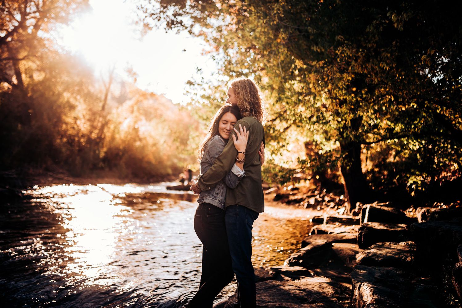 Couple embracing by a sunlit stream with golden fall foliage in background.