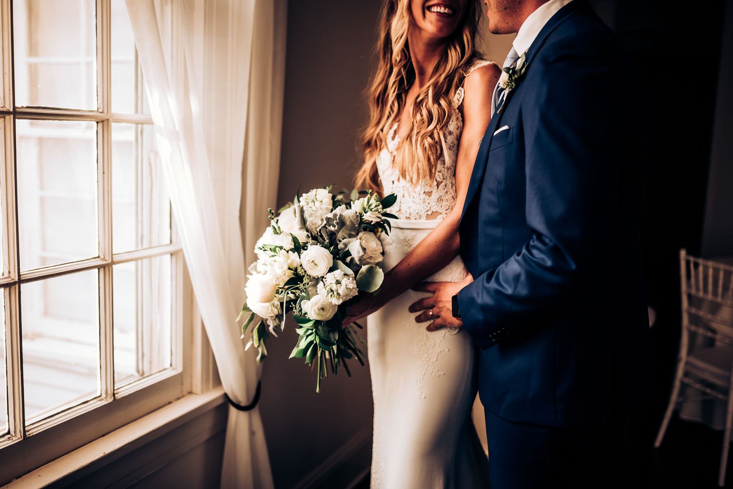 Sunlit wedding moment by window featuring white bouquet and navy suit details.