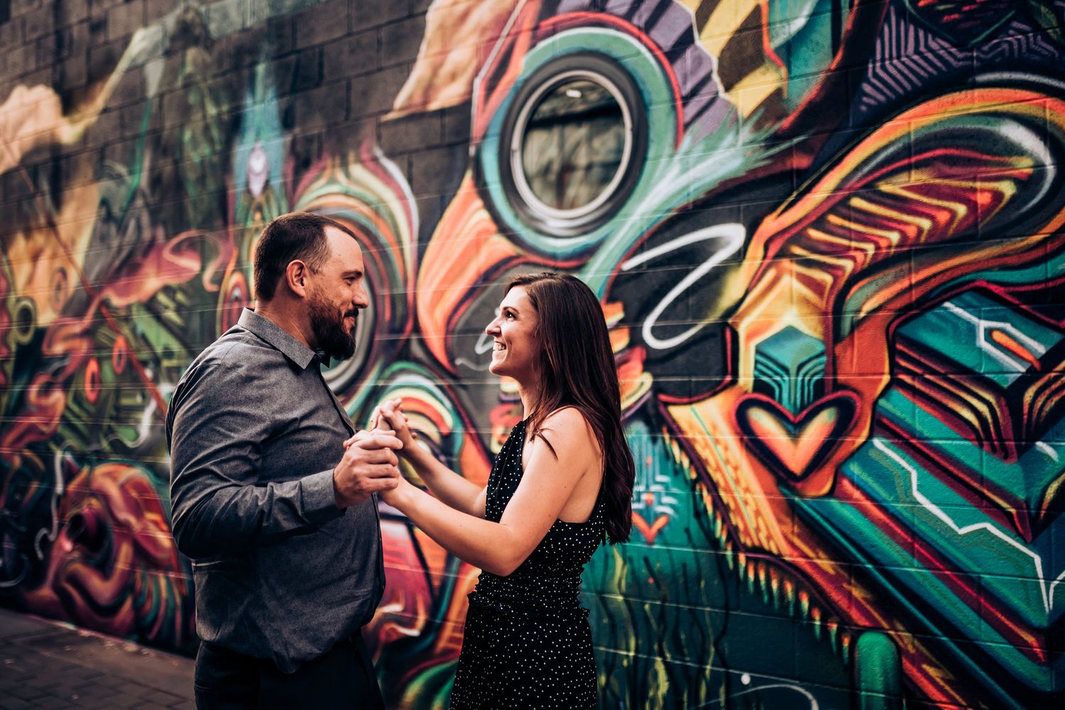 A couple shares a joyful moment against a wall with colorful street art featuring swirling designs.