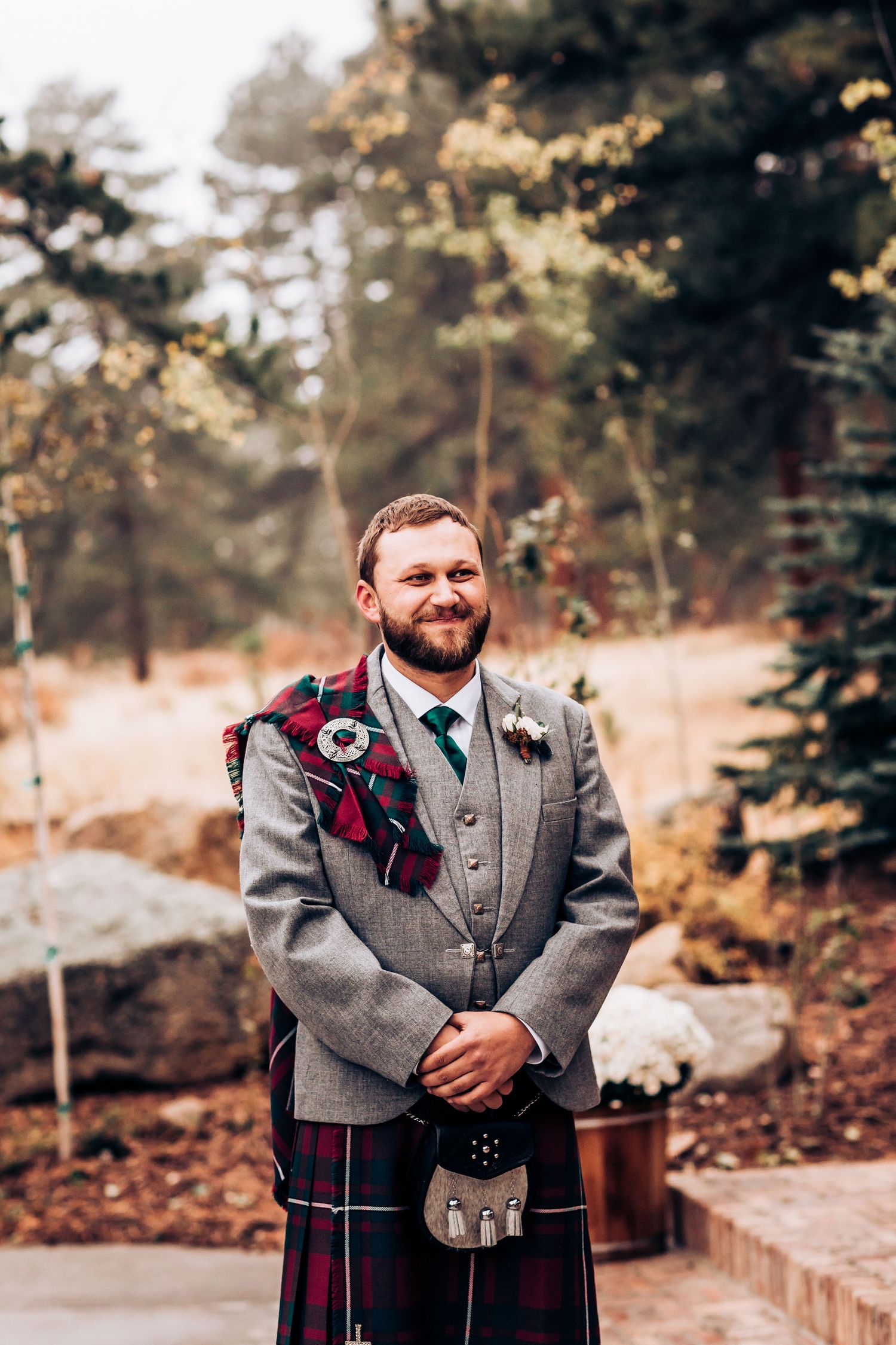 A groom wearing a traditional Scottish kilt and gray tweed jacket smiles outdoors during an autumn wedding ceremony.