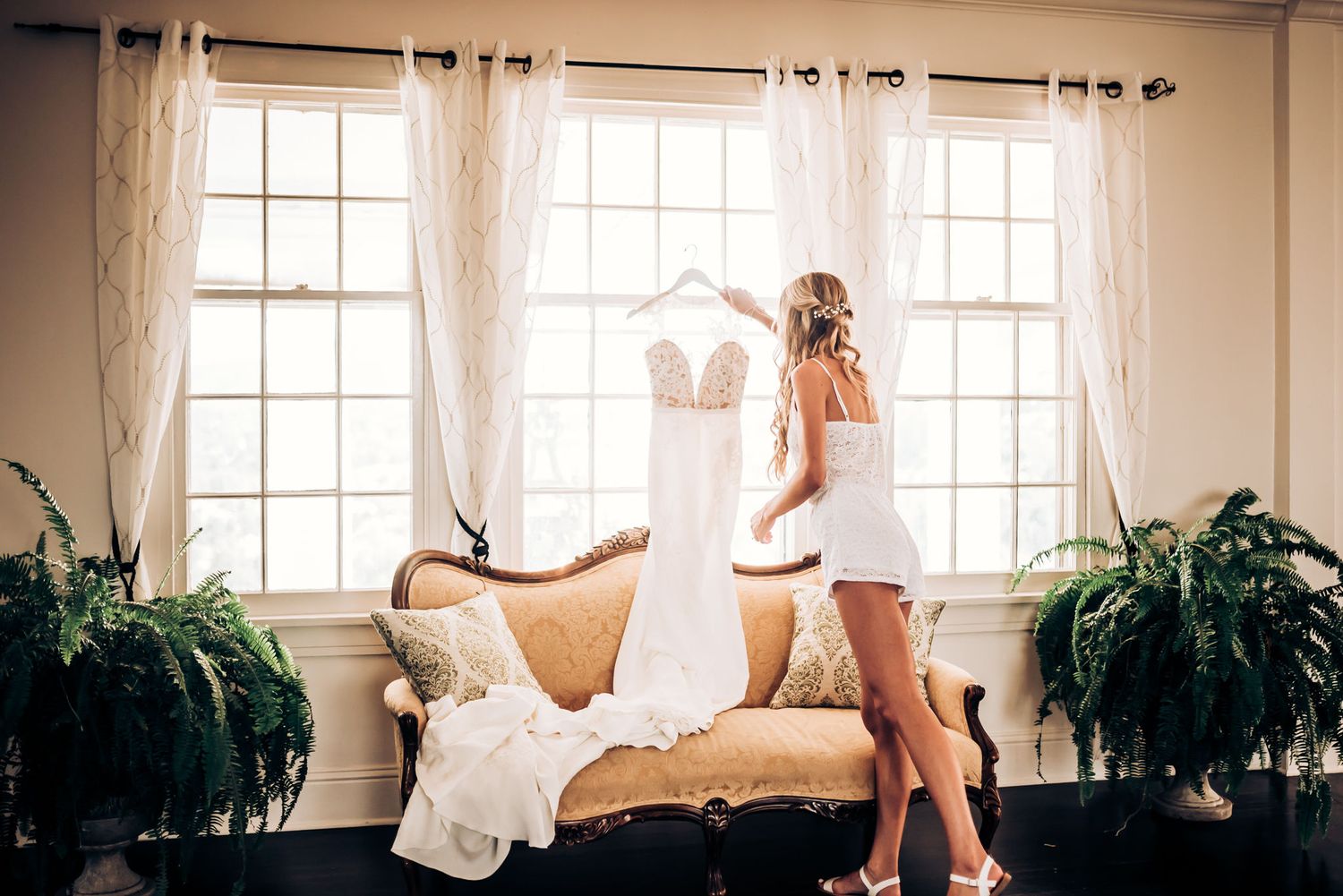 Woman in white dress looking at wedding gown hanging by sunny windows with indoor plants.