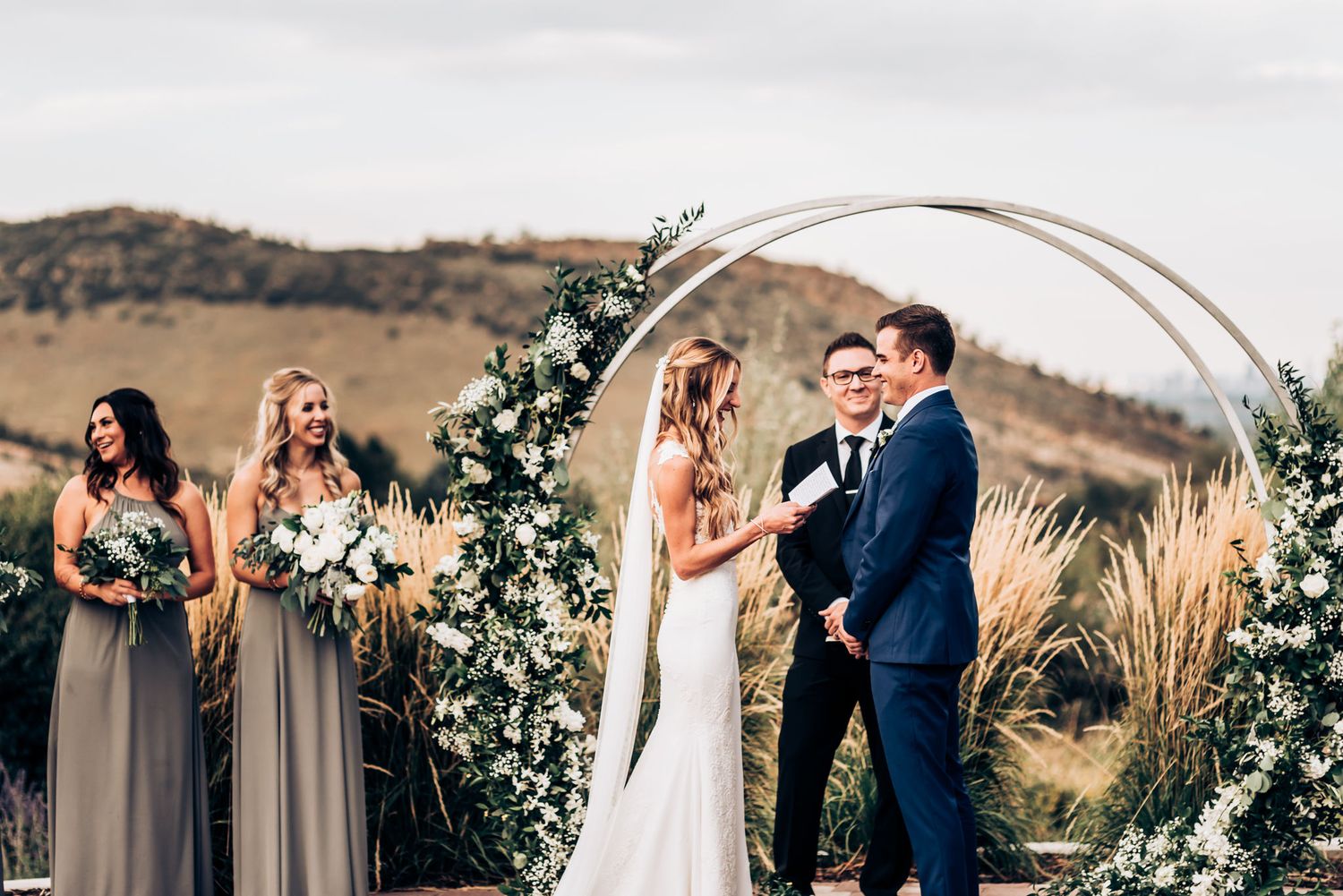 Outdoor wedding ceremony under circular arch decorated with white flowers and greenery against mountain backdrop.