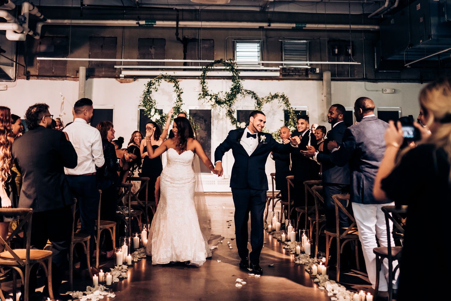 Newlyweds exit down candlelit aisle in industrial venue while wedding guests cheer during recessional.