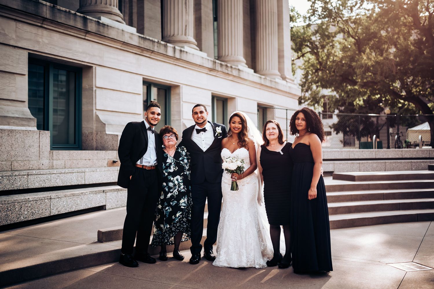 A wedding party poses on grand stone steps outside a classical building.