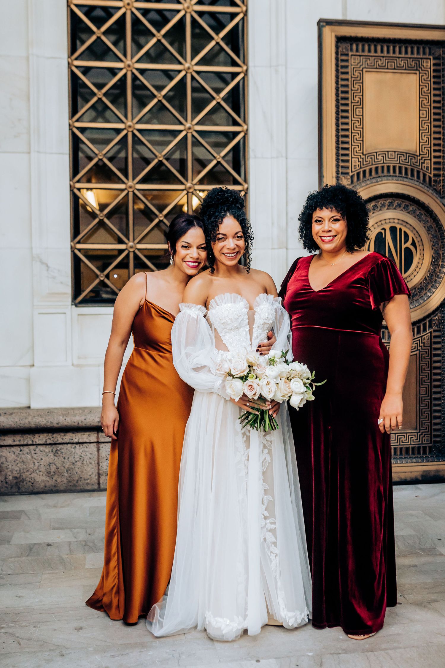 Wedding party photo with bride in white and attendants in burgundy and rust-colored gowns.