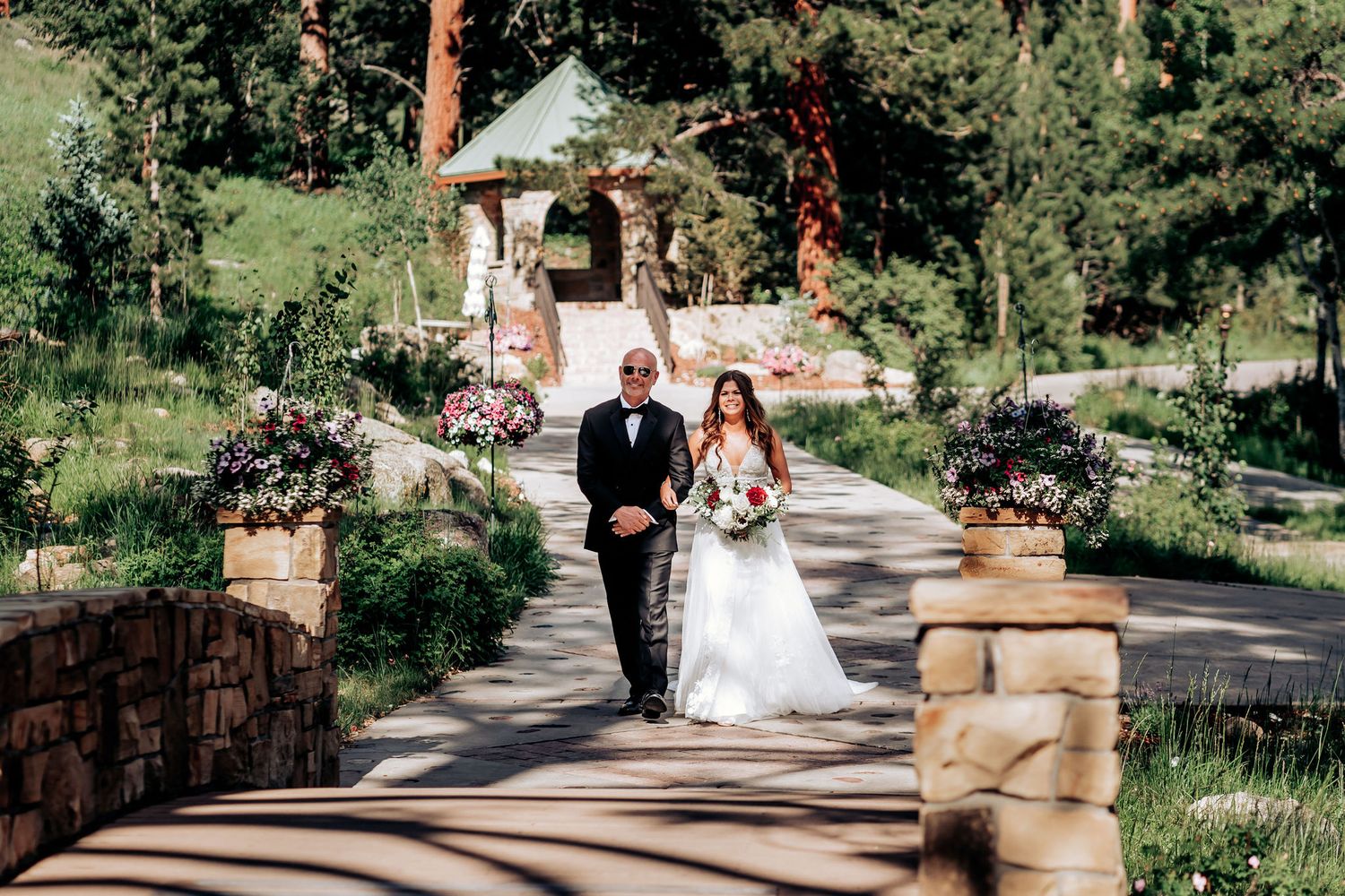 A couple walks down a scenic stone pathway surrounded by lush greenery and pine trees.