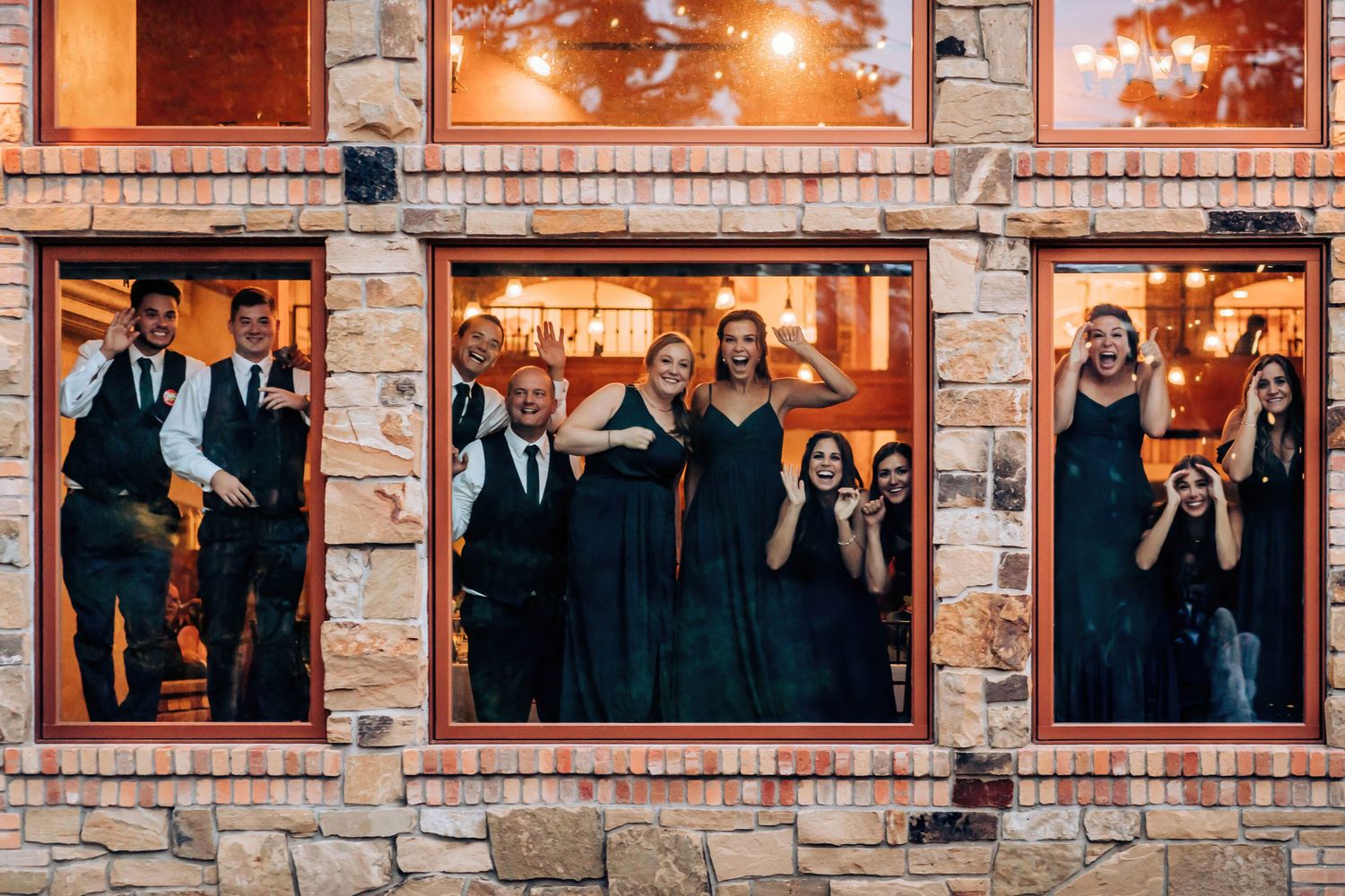 Guests in formal attire pose through window frames of a brick building during an evening celebration.