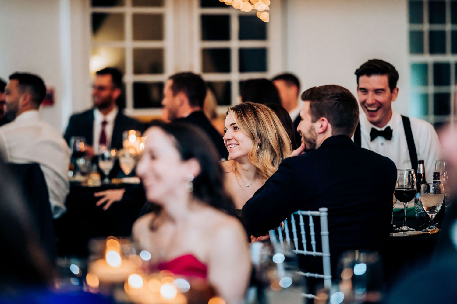 Wedding reception guests laugh and socialize at elegantly set tables with glowing candlelight.