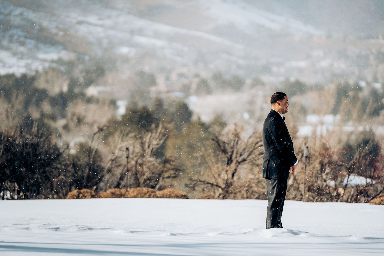 A solitary figure stands in a black suit on a snow-covered hill overlooking a distant mountain landscape.