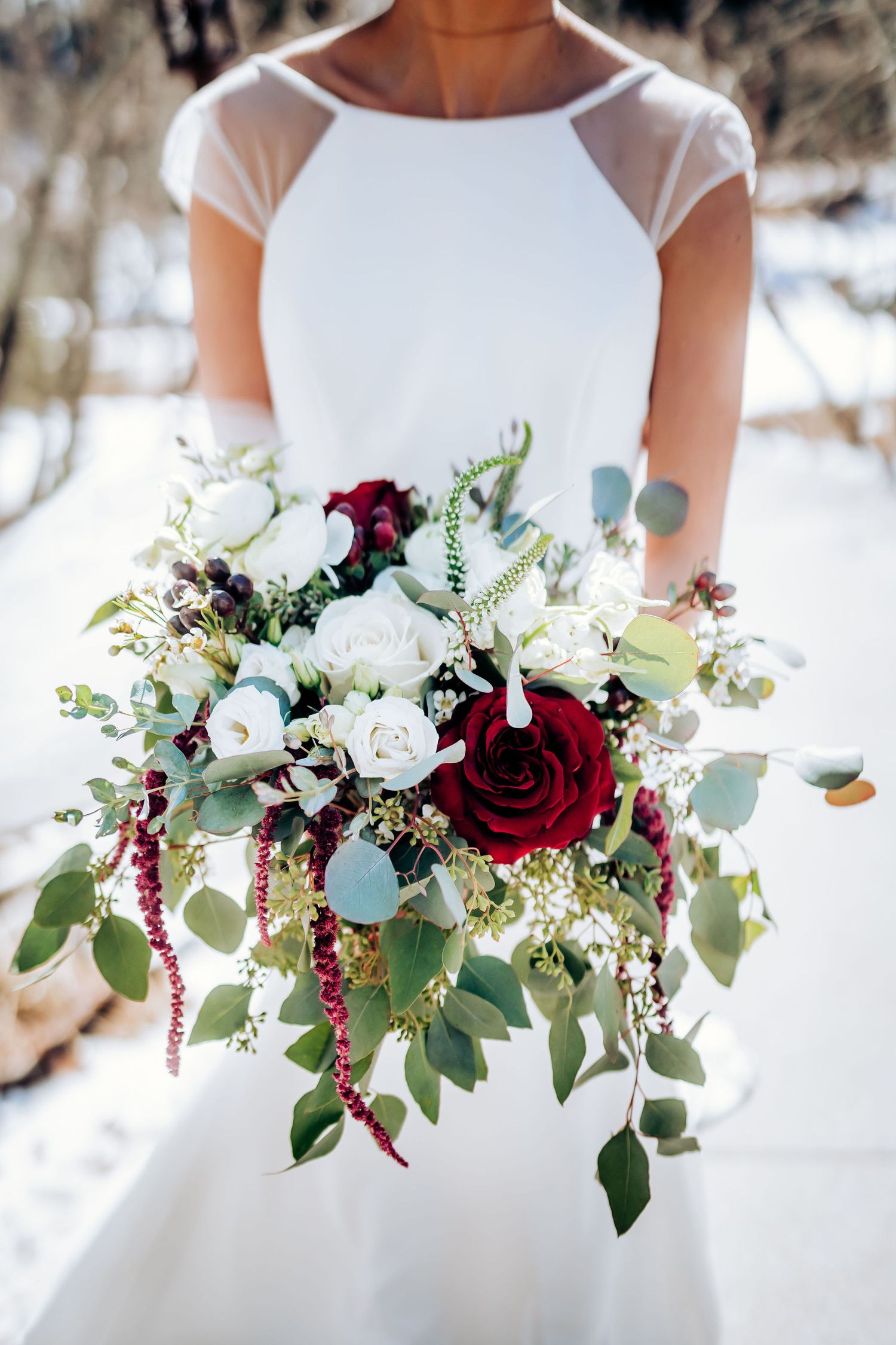 Elegant bridal bouquet featuring deep red roses, white flowers, and cascading eucalyptus leaves against snow.