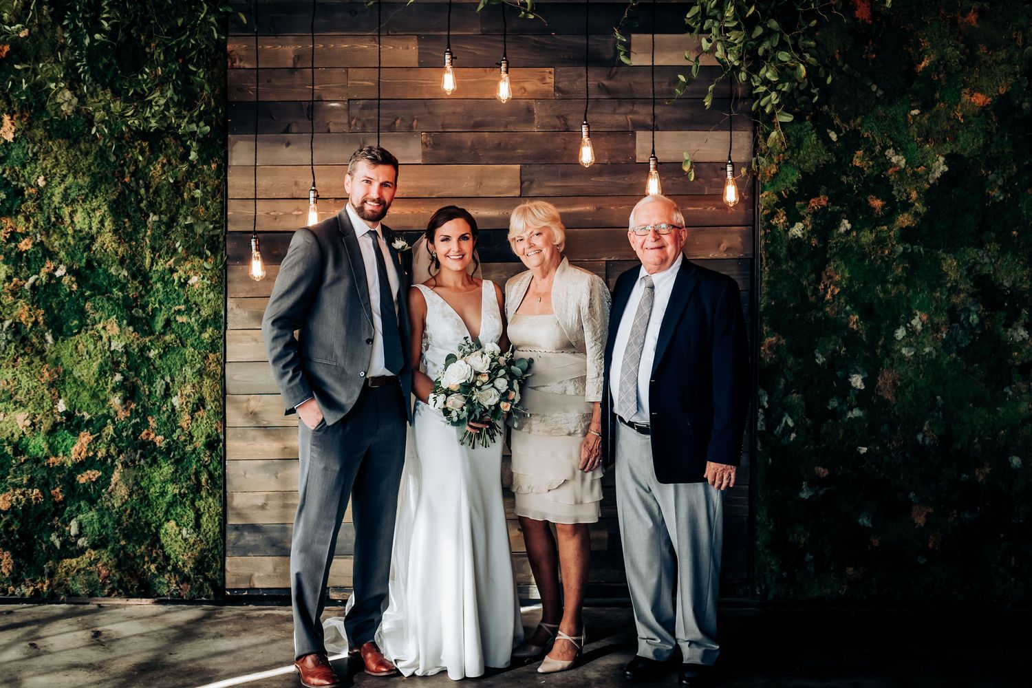 Wedding portrait against rustic wooden wall with pendant lights and climbing vines.