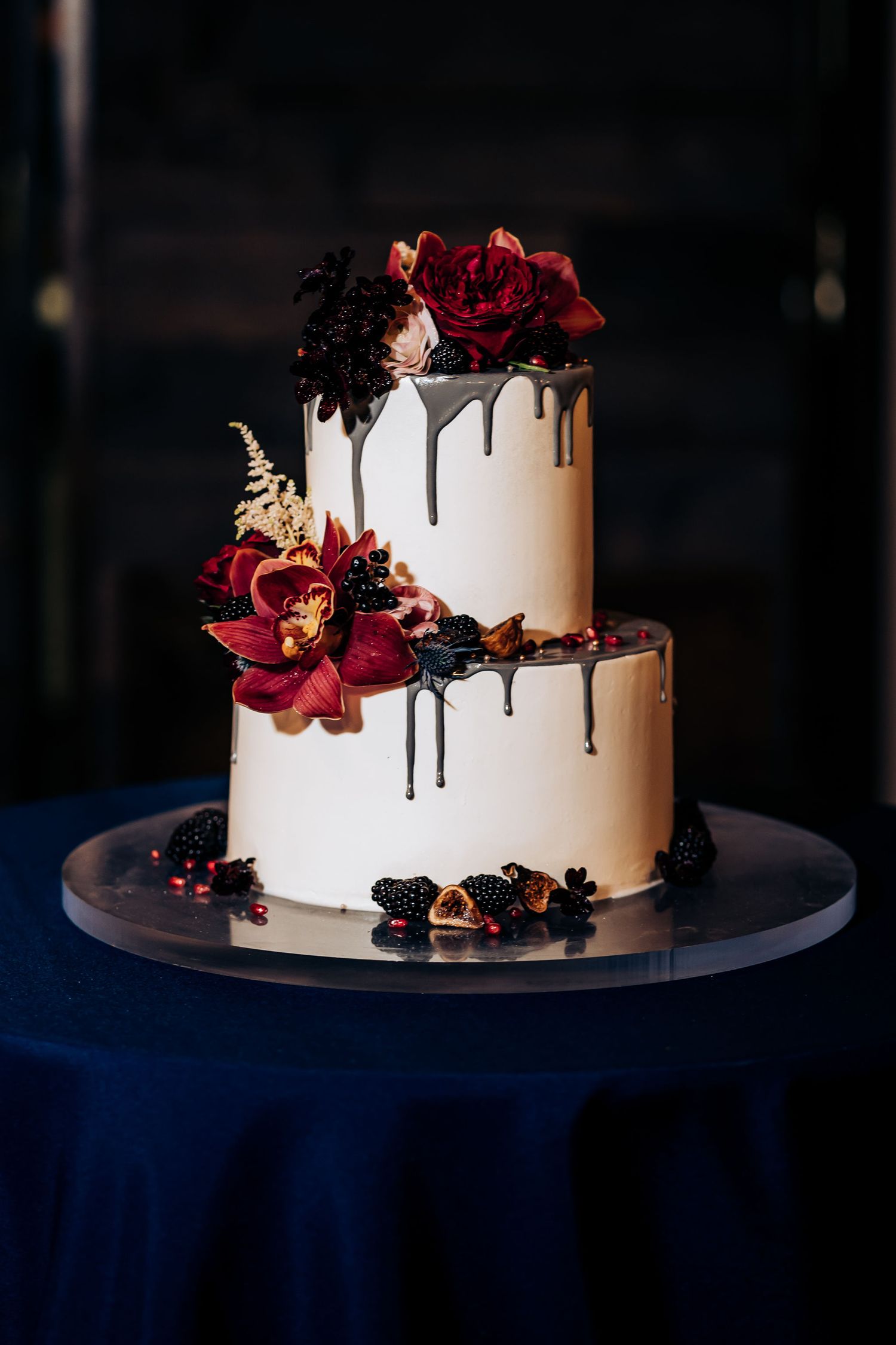 Elegant two-tier white wedding cake with dark drips and red floral decorations on navy tablecloth.